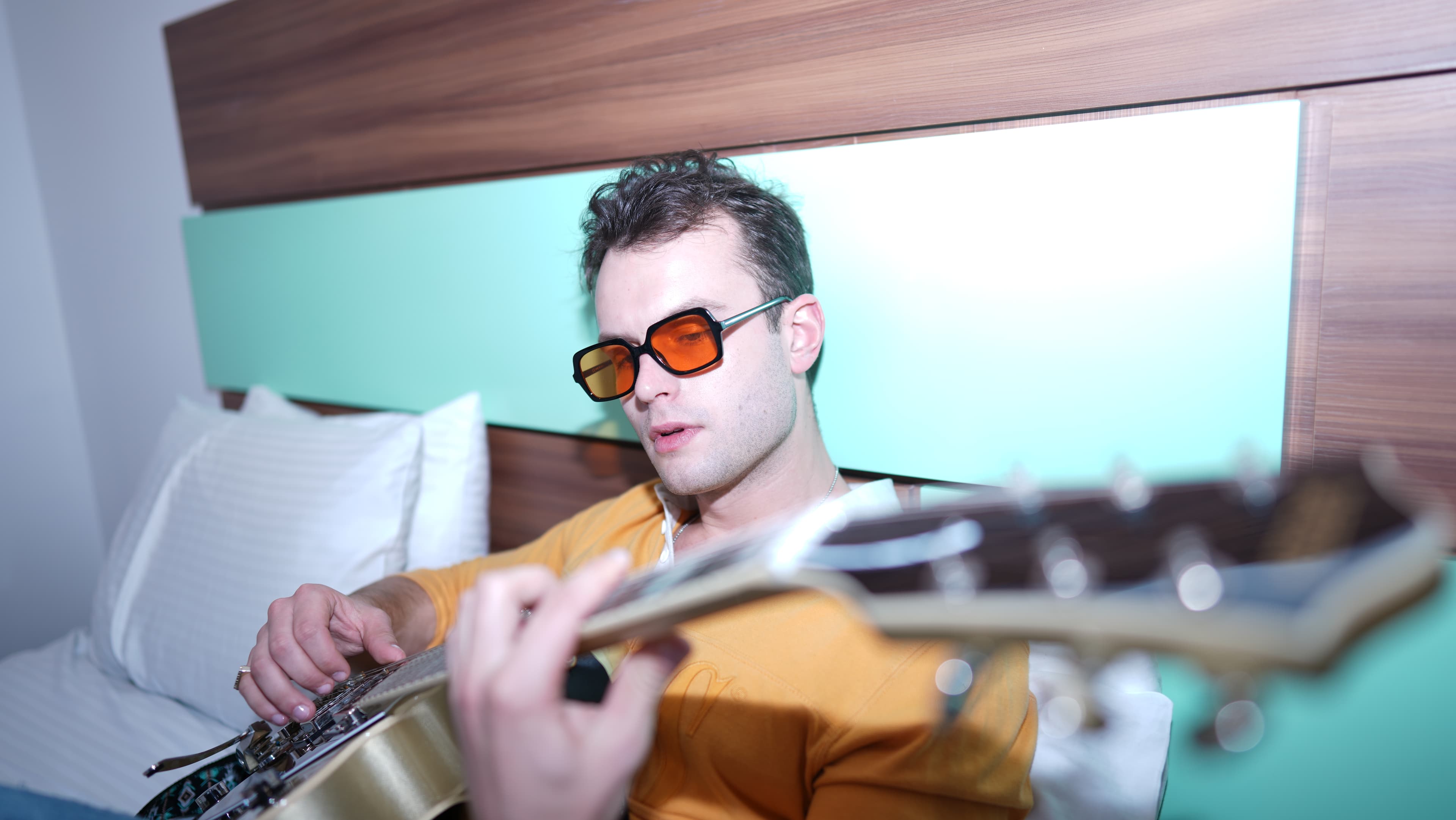 Sébastien Graux joue de la guitare sur un lit d'hôtel — le genre de session où une guitare à 99$ et un iPhone sont devenus un placement Ricky Martin